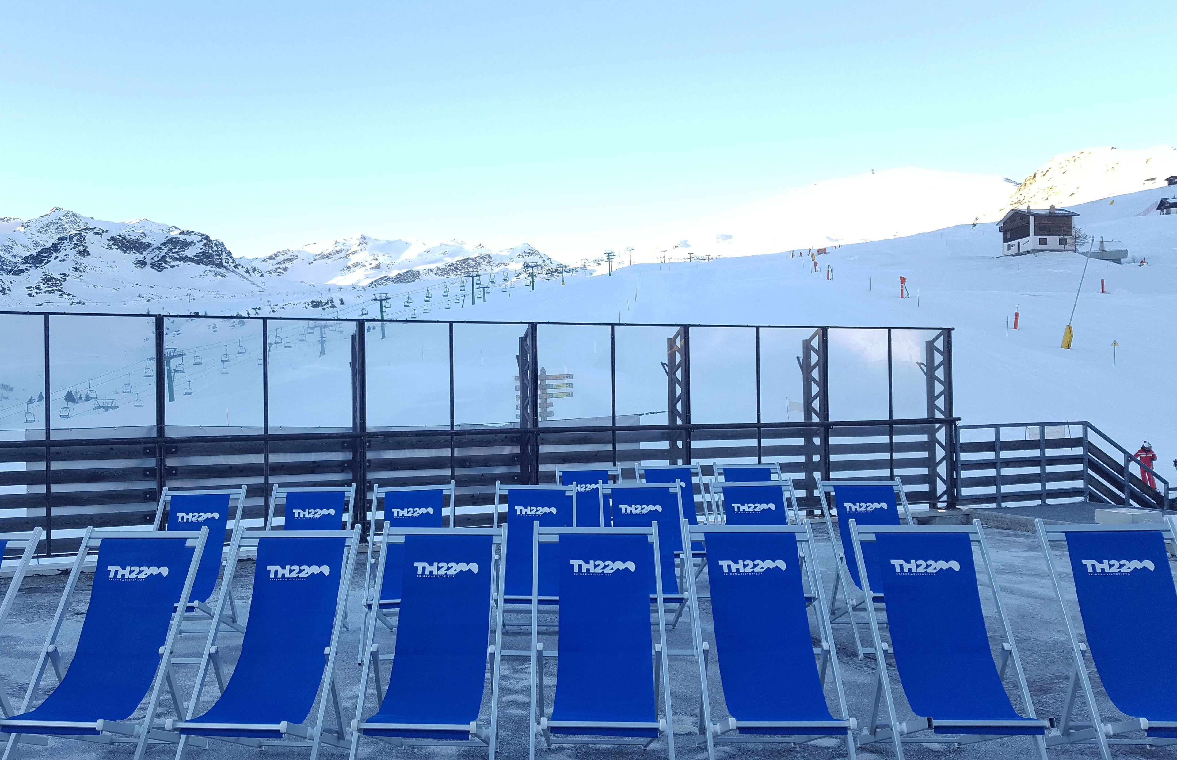 A ski lift area with blue chairs labeled TH22M, a wooden cabin, and a snow-covered mountain backdrop.