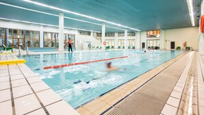 Ein Hallenbad mit Bahnen, einem Rettungsschwimmer und einer Treppe, die zu einer oberen Ebene führt.