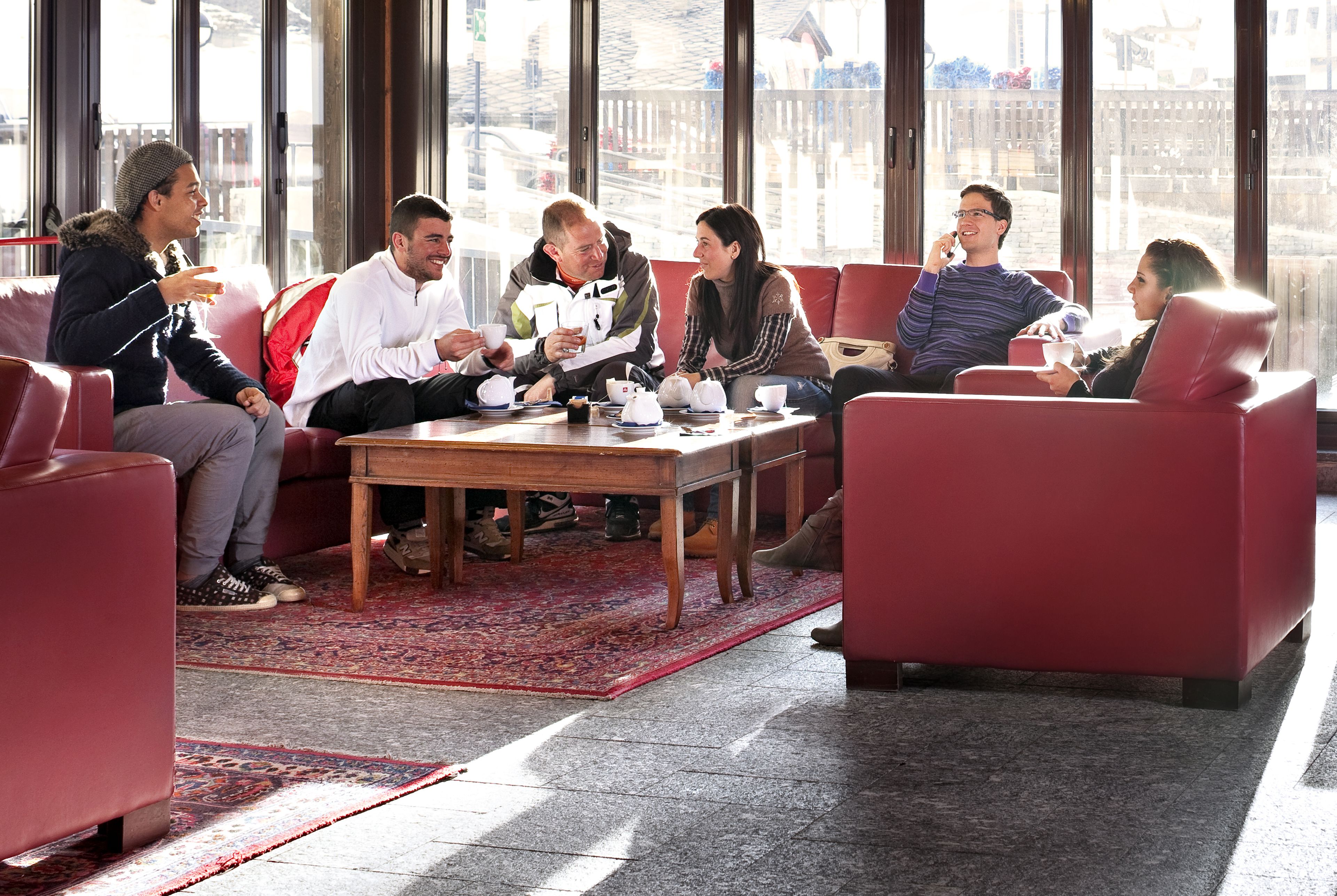A group of people seated on red couches in a room with large windows, a wooden table, and a patterned rug.
