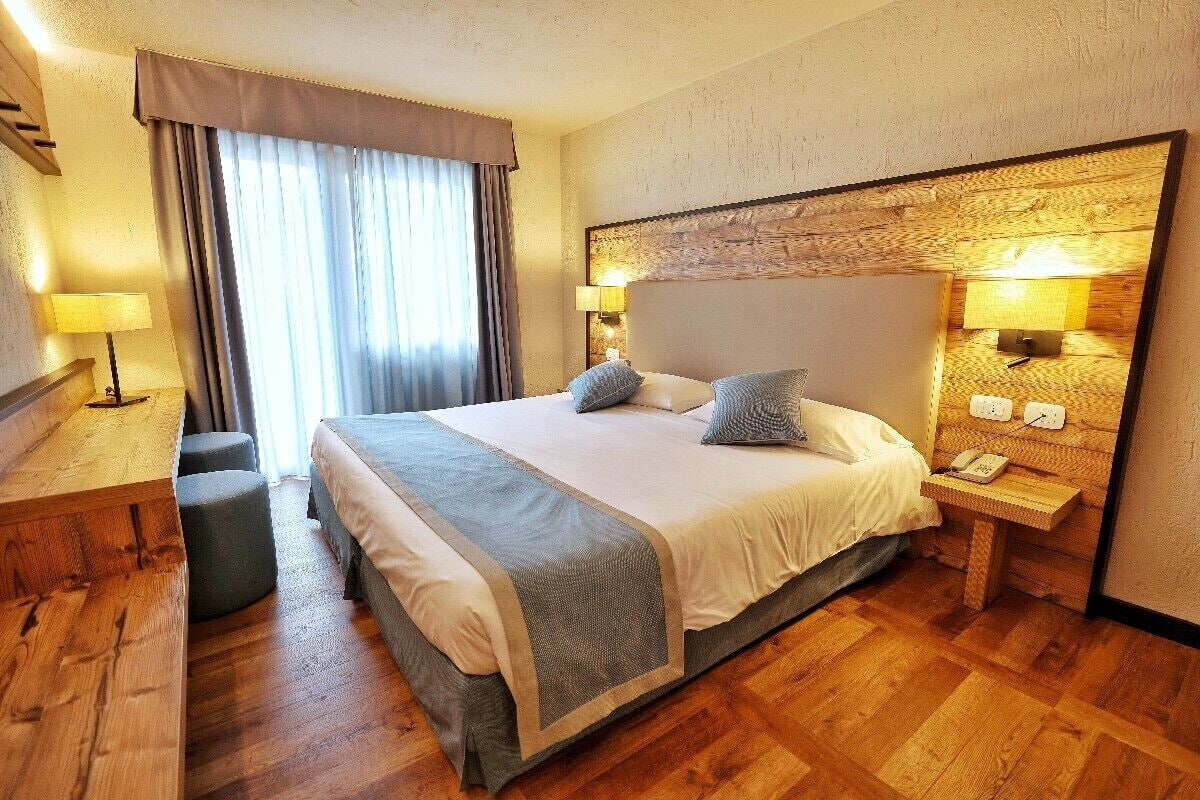 Ein Hotelzimmer mit einem großen Bett, zwei Nachttischen mit Lampen, einem Holztisch und einem großen Fenster mit Vorhängen.