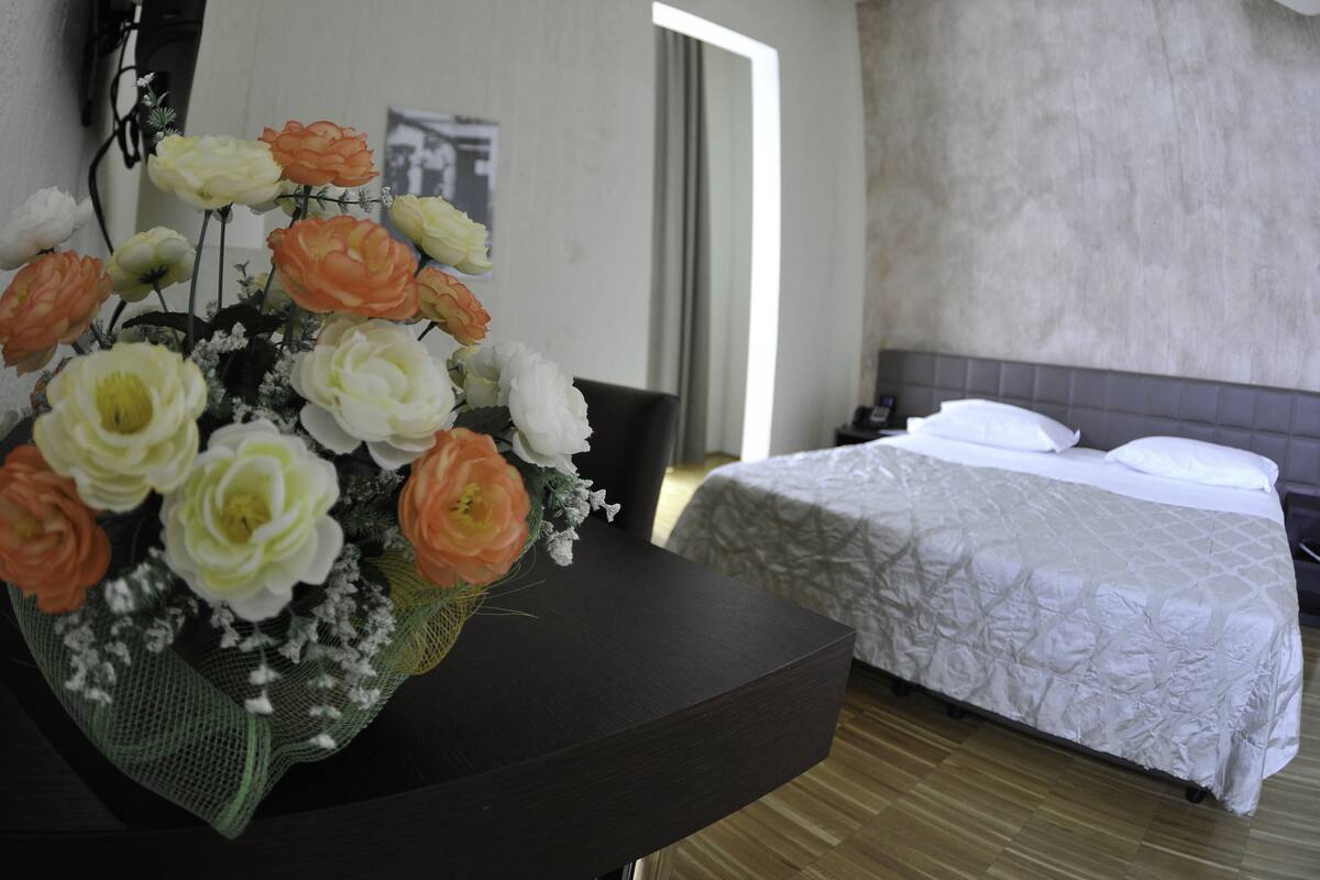 A hotel room with a bed, a nightstand, and a desk with a bouquet of flowers.