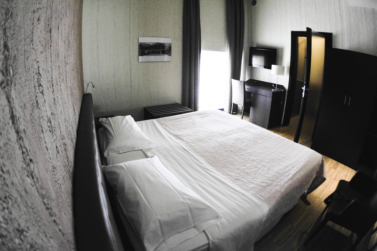 A hotel room with a large bed, a desk, a chair, a TV, and a wardrobe.