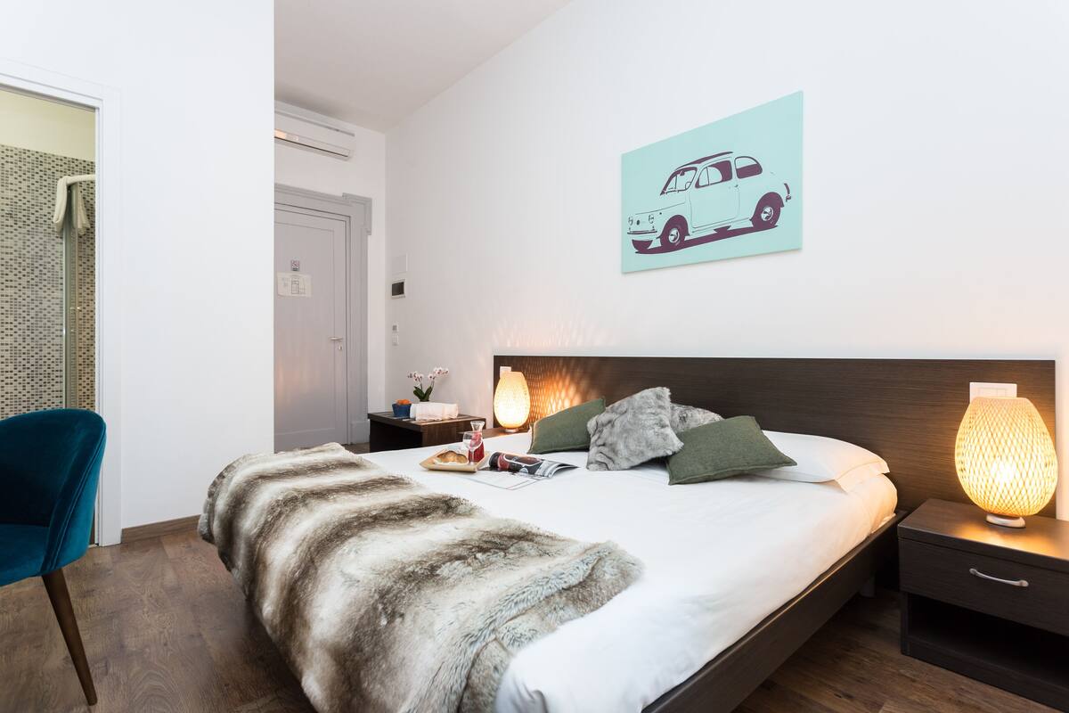 Een keurig opgemaakt slaapkamer met een bed, nachtkastje, lamp, stoel en een kunstwerk aan de muur.
