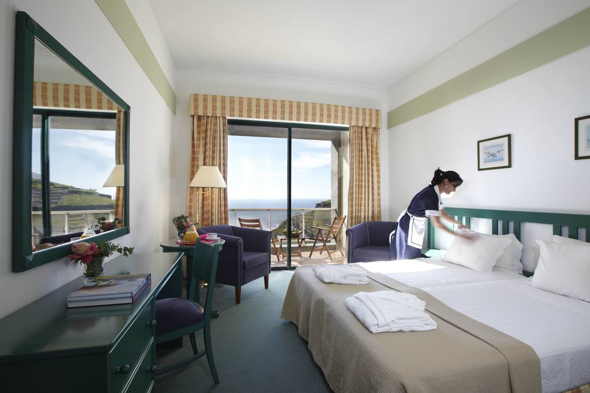 A hotel room with a large bed, a desk with a chair, a small table with a vase of flowers, and a view of the ocean through a large window.
