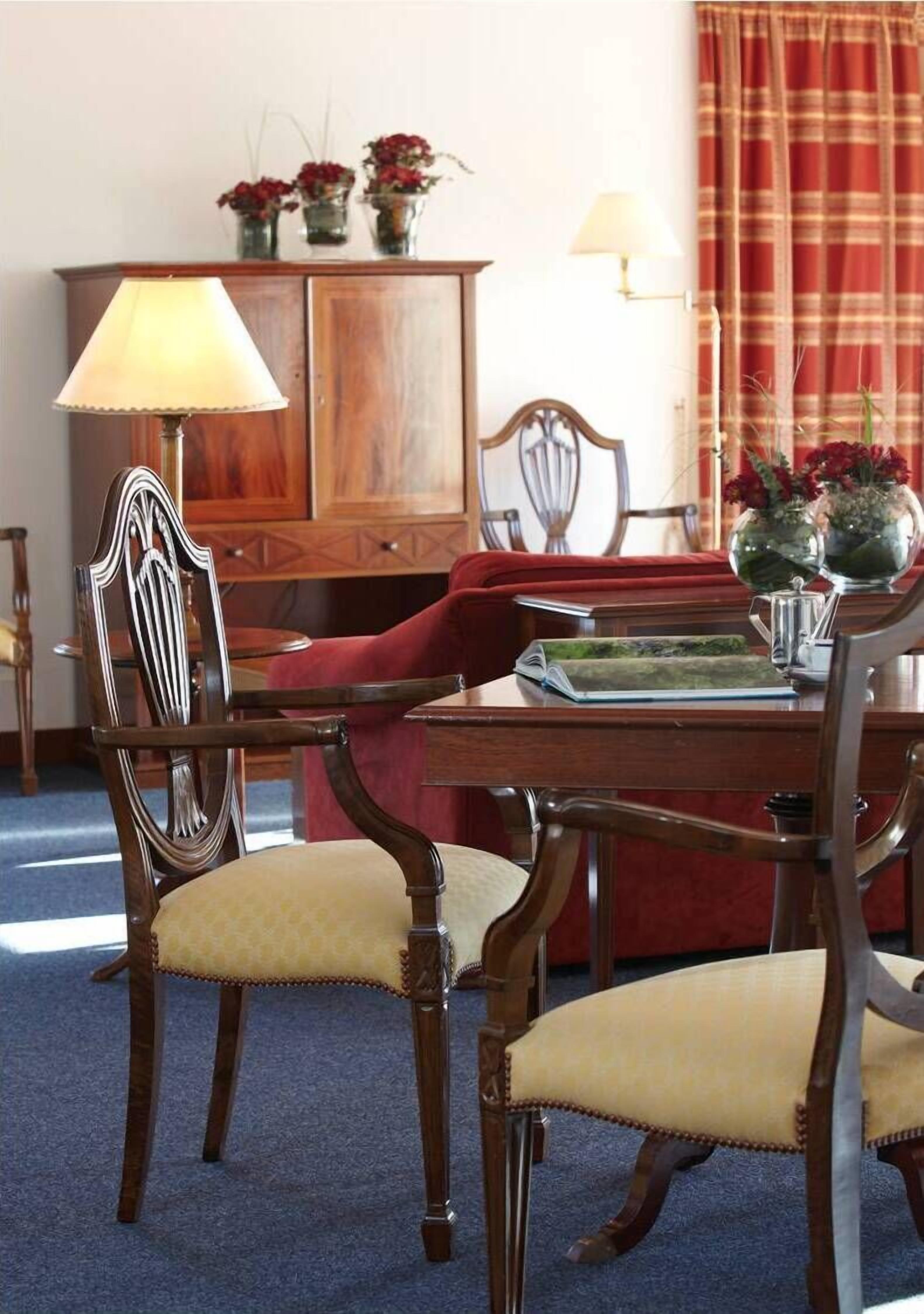 Un coin repas comprenant une table en bois, quatre chaises, un banc recouvert d’un tissu rouge, un buffet et une lampe.