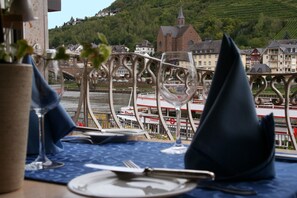 A dining table set with a wine glass, a folded napkin, and a plate, overlooking a river with boats and buildings in the background. - Hotel Karl Noss (Cochem)