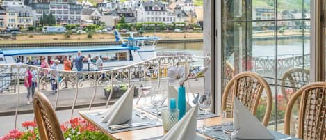 A dining area with tables set for two, overlooking a river with boats and a town in the background.
