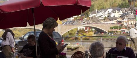 An outdoor café with red umbrellas, wicker chairs, and a view of a town with a bridge.