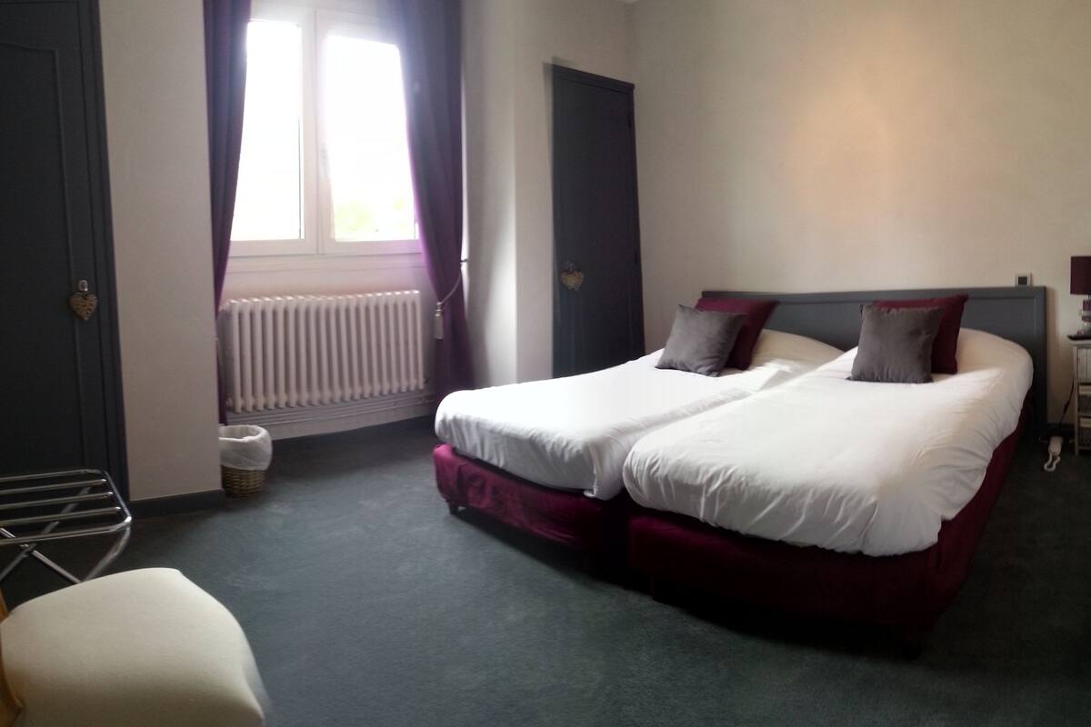 Quarto de hotel com duas camas, uma cadeira, um radiador e uma pequena mesa com abajur.