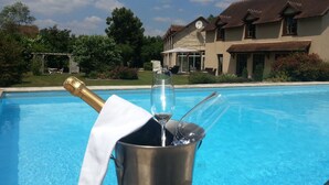 A swimming pool with a champagne bottle and glasses in a bucket, surrounded by a house with a tiled roof and a garden.