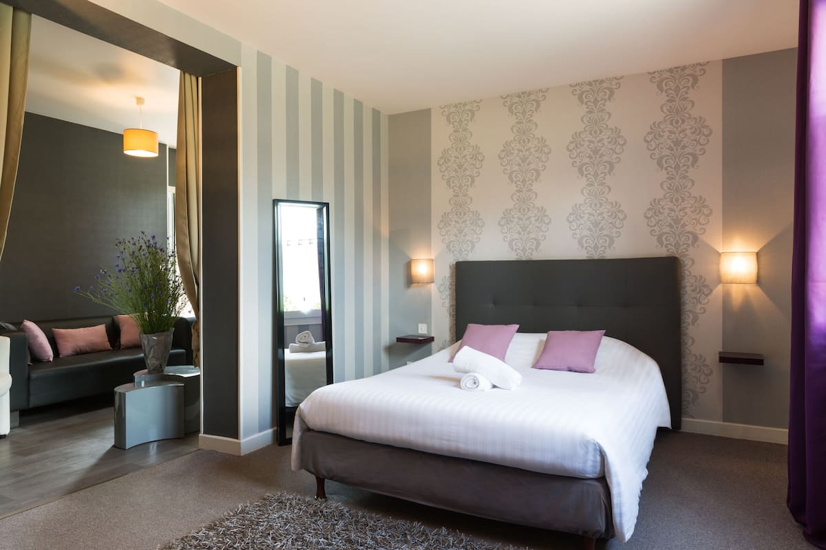 Quarto de hotel moderno com uma cama grande, mesas de cabeceira, um espelho e um espaço de estar com um sofá e uma mesa pequena.