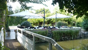 A wooden bridge over a calm water body, leading to an outdoor seating area with umbrellas and tables, surrounded by lush greenery.
