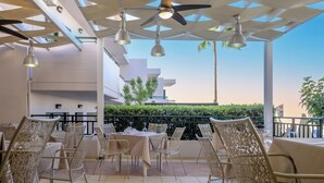 Un área de estar al aire libre con mesas y sillas, un ventilador de techo y vistas a un edificio y vegetación.