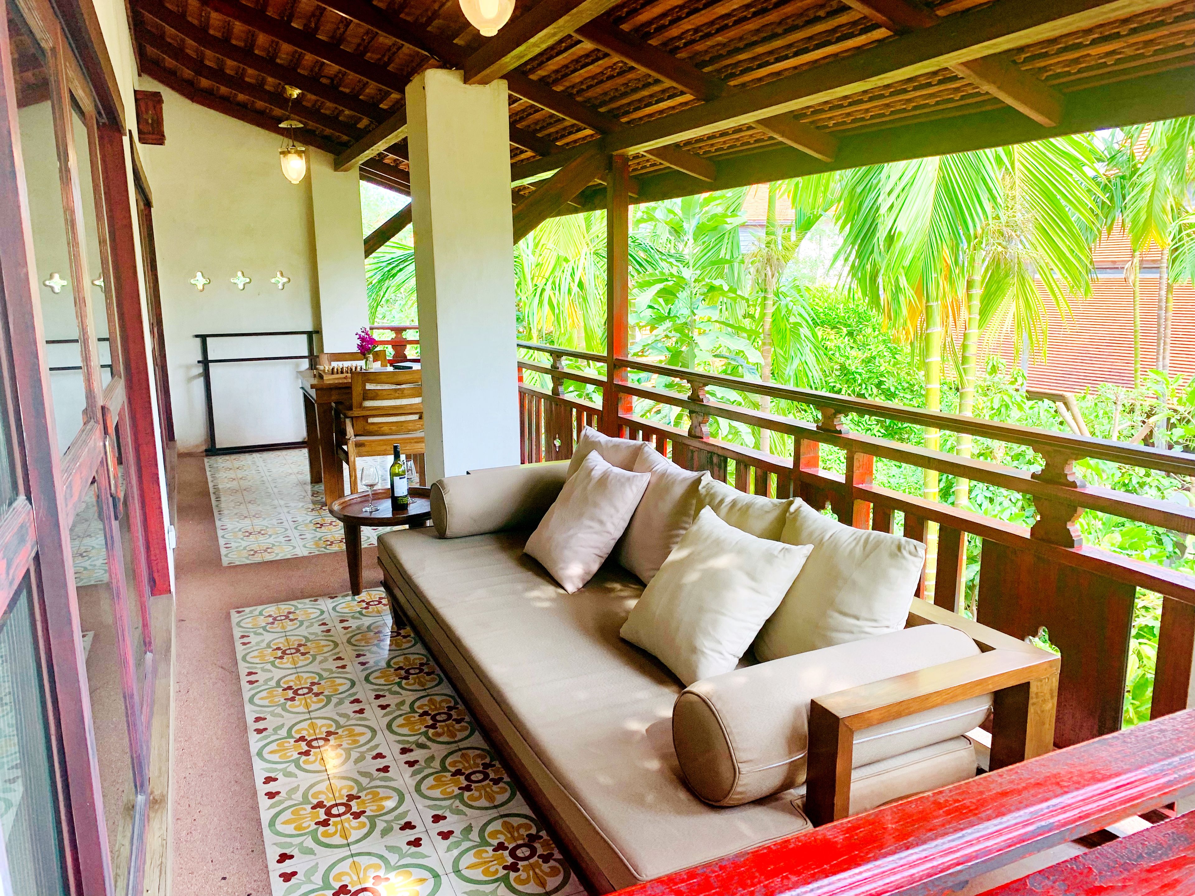 A covered outdoor seating area with a sofa and a table, surrounded by a wooden railing with a view of lush greenery.