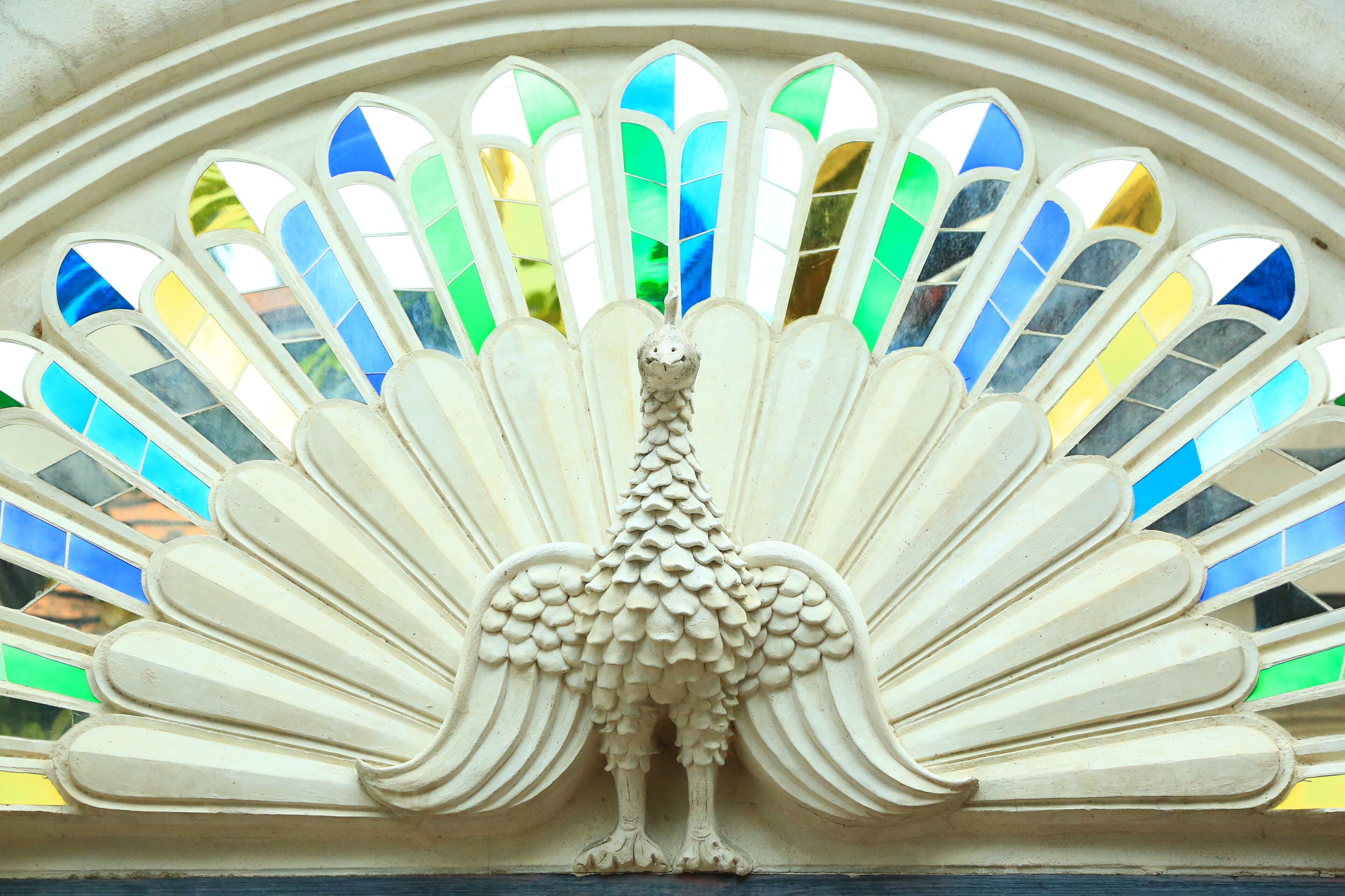 A detailed architectural carving of a peacock with a fan of colorful stained glass windows behind it.