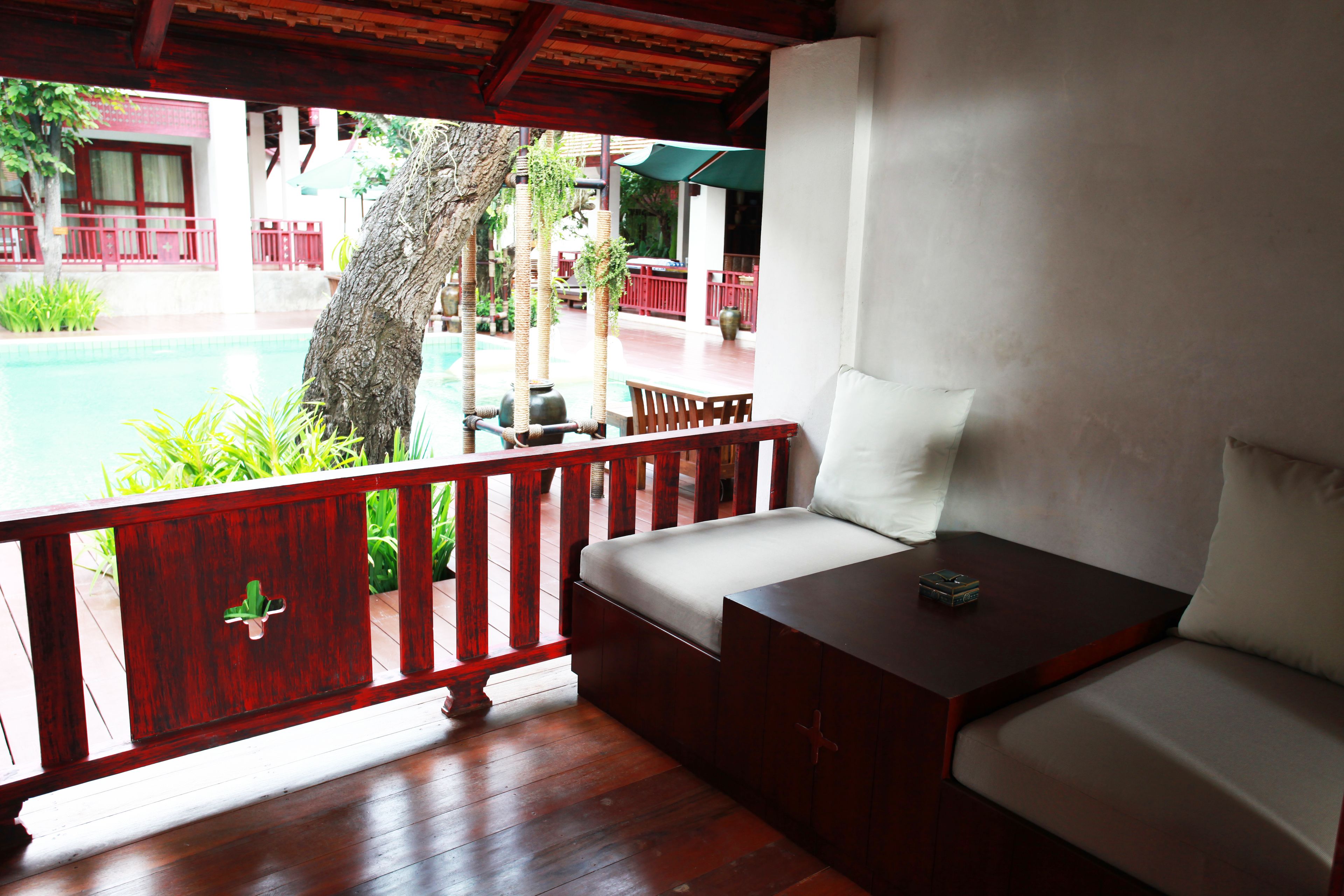 A wooden deck with a sofa and a table, overlooking a pool and a building with red railings.