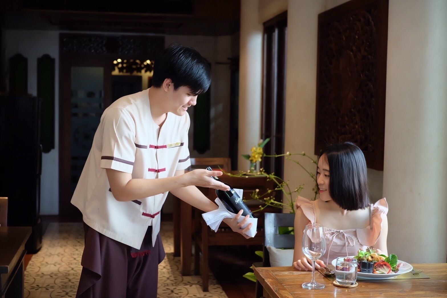 A person serving food to a guest at a table in a restaurant with wooden furniture and a decorative wall.
