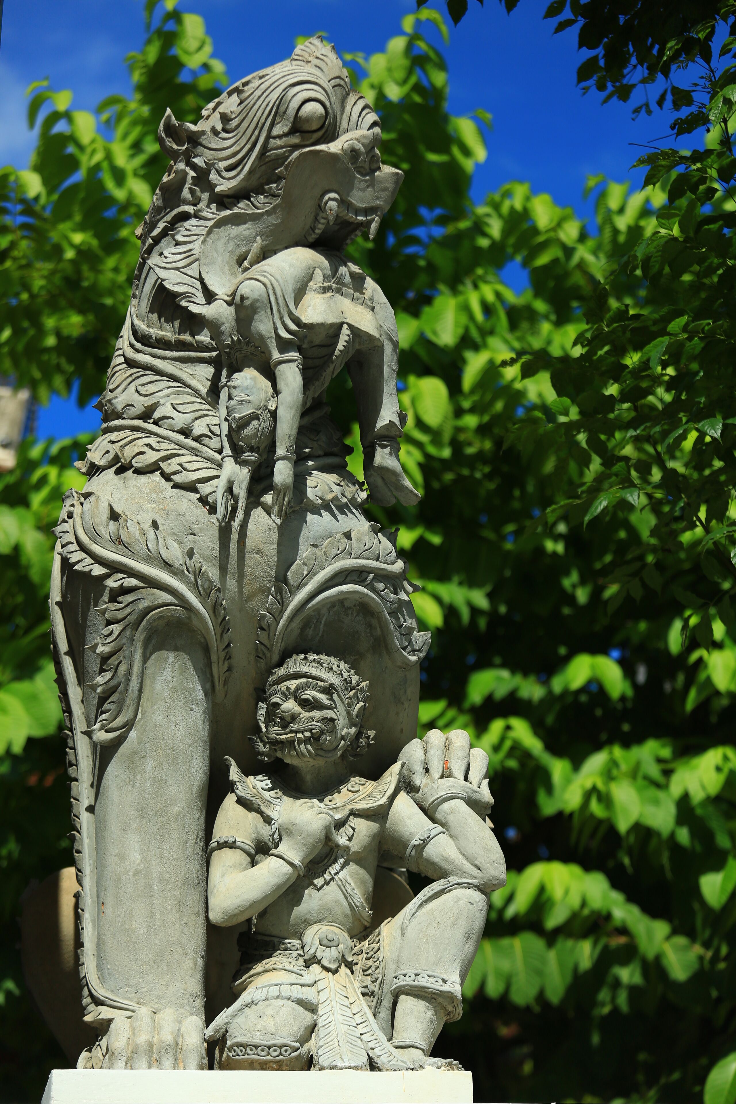 A detailed stone statue with intricate carvings, set against a backdrop of lush green foliage.