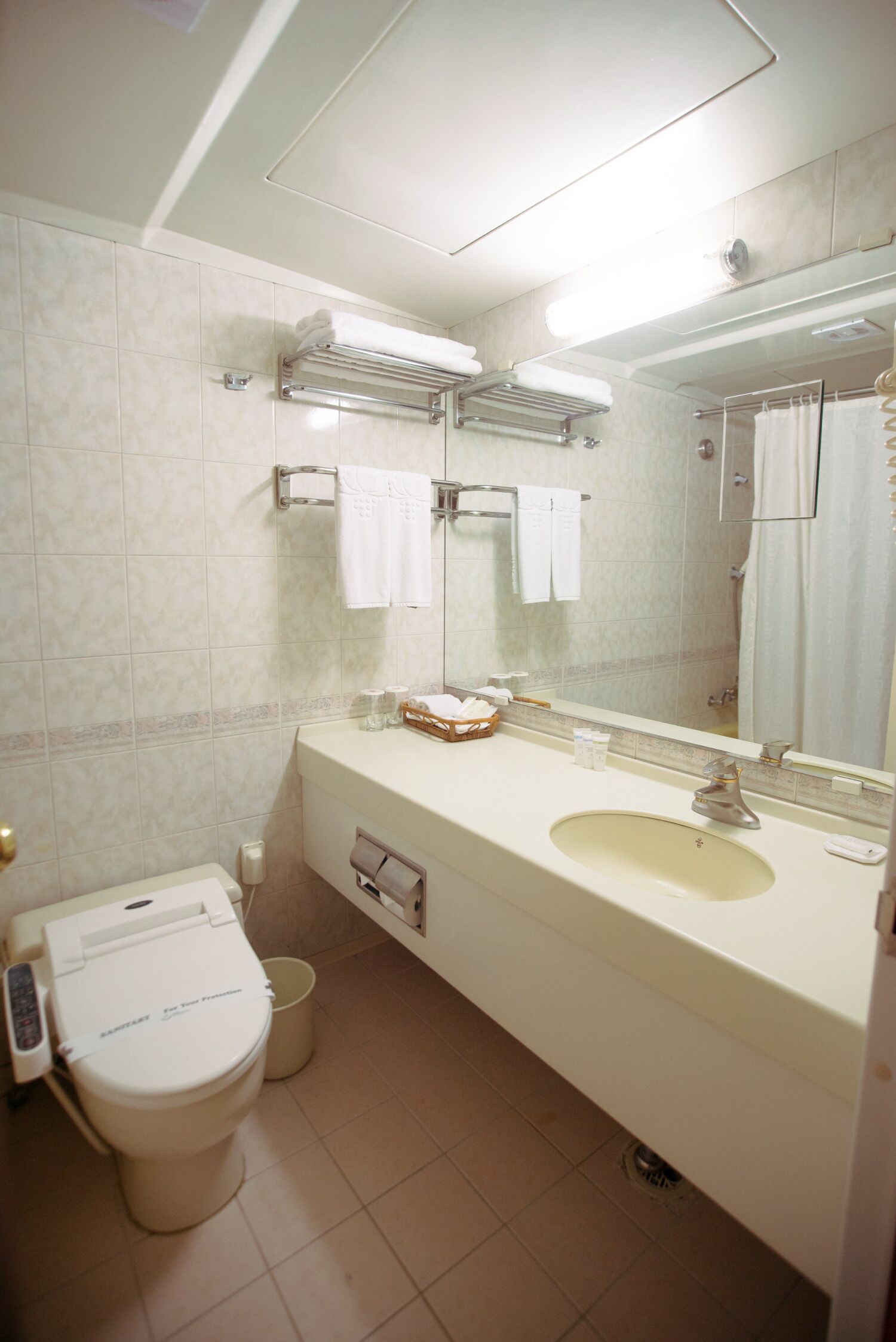 Un baño con inodoro, lavamanos y espejo. Hay toallas colgadas en un perchero, una cortina de ducha y un secatoallas.