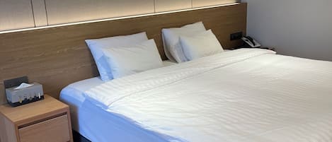Un lit bien fait, avec du linge de lit blanc, une table de chevet en bois sur laquelle se trouve une boîte de mouchoirs, et un téléphone fixé au mur.