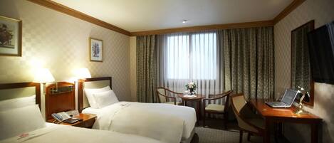 A hotel room with two beds, a desk with a laptop, a chair, a small table with a vase, and curtains on the window.