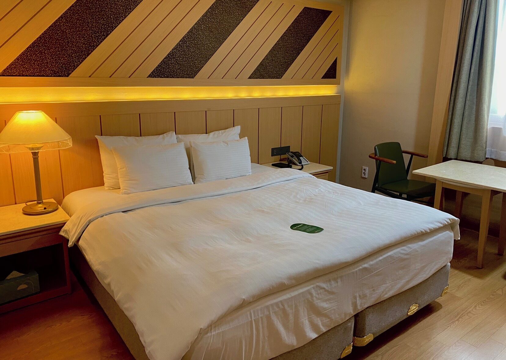 Una habitación de hotel con una cama, una mesita de noche con lámpara, una silla, una pequeña mesa y un teléfono en el buró.