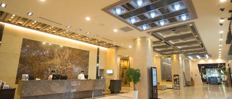 A modern hotel lobby with a marble reception desk, a large potted plant, and a high ceiling with recessed lighting.