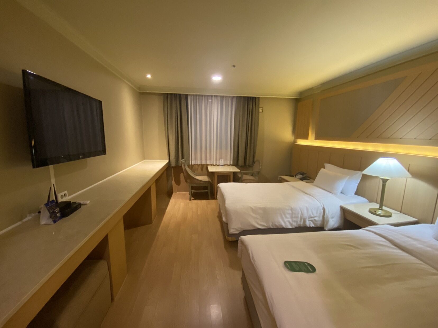 A hotel room with two beds, a desk, a chair, a TV, and a lamp.
