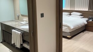 A modern bedroom with a large bed, a bedside table, and a nightstand. There is a bathroom with a sink, mirror, and towel hanging on the door. - Hotel International (Changwon)