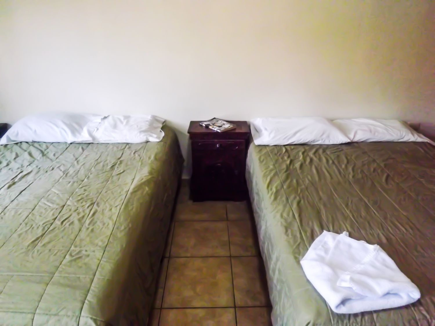 Quarto com duas camas de solteiro, cada uma com colcha verde e almofadas brancas. Há uma pequena mesa de cabeceira de madeira entre as camas, com um objeto de cor escura sobre ela.