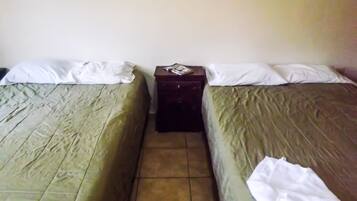 Quarto com duas camas de solteiro, cada uma com colcha verde e almofadas brancas. Há uma pequena mesa de cabeceira de madeira entre as camas, com um objeto de cor escura sobre ela.