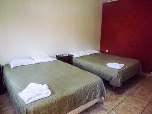 Una habitación con dos camas individuales, cada una con una colcha verde y almohadas blancas. Entre las camas hay una mesita de noche de madera con una lámpara encima. Las paredes son lisas, y una de ellas está pintada de rojo.
