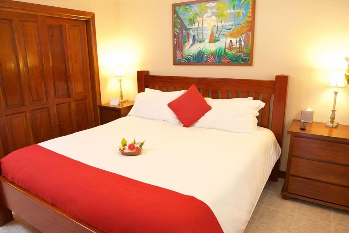A neatly made bed with a red and white bedspread, a red pillow, and a small fruit bowl on the bed. There are two bedside tables with lamps, a wooden wardrobe, and a painting on the wall depicting a tropical scene.