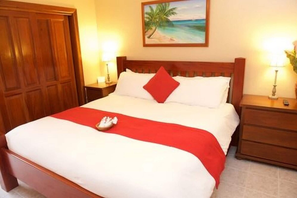 Habitación de hotel con cama, cabecera de madera, almohada roja, mesita de noche con lámpara y un cuadro de una playa.