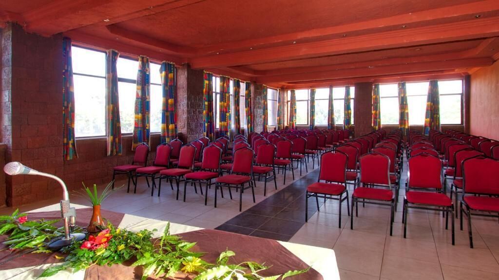A conference room with rows of red chairs, large windows with colorful curtains, and a potted plant on the floor.