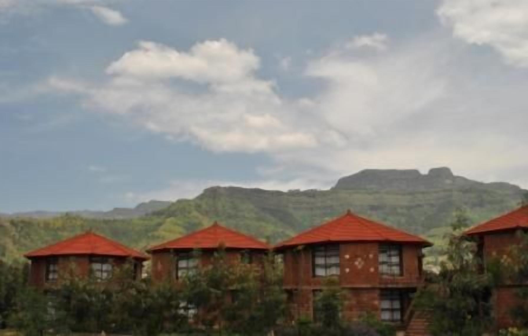 A row of four brick buildings with red roofs, set against a backdrop of green hills and a cloudy sky.
