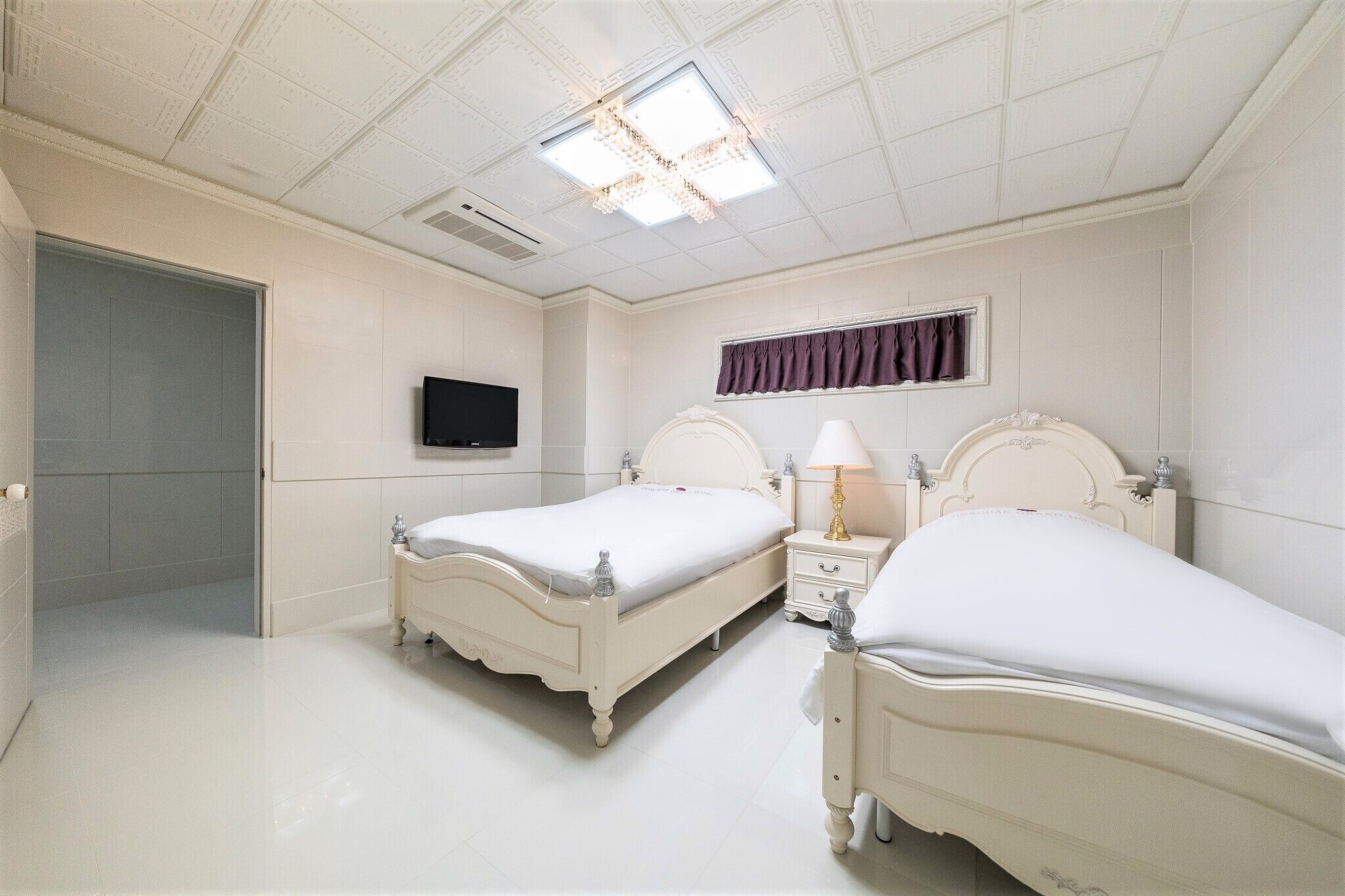 A bedroom with two white beds, a TV, a window with curtains, and a ceiling light fixture.