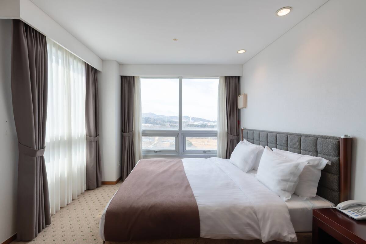 Ein Hotelzimmer mit einem großen Bett, einem Nachttischtelefon und Blick auf die Stadt durch das Fenster.