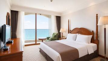 A hotel room with a bed, a desk with a television, a chair, a lamp, and a view of the sea.