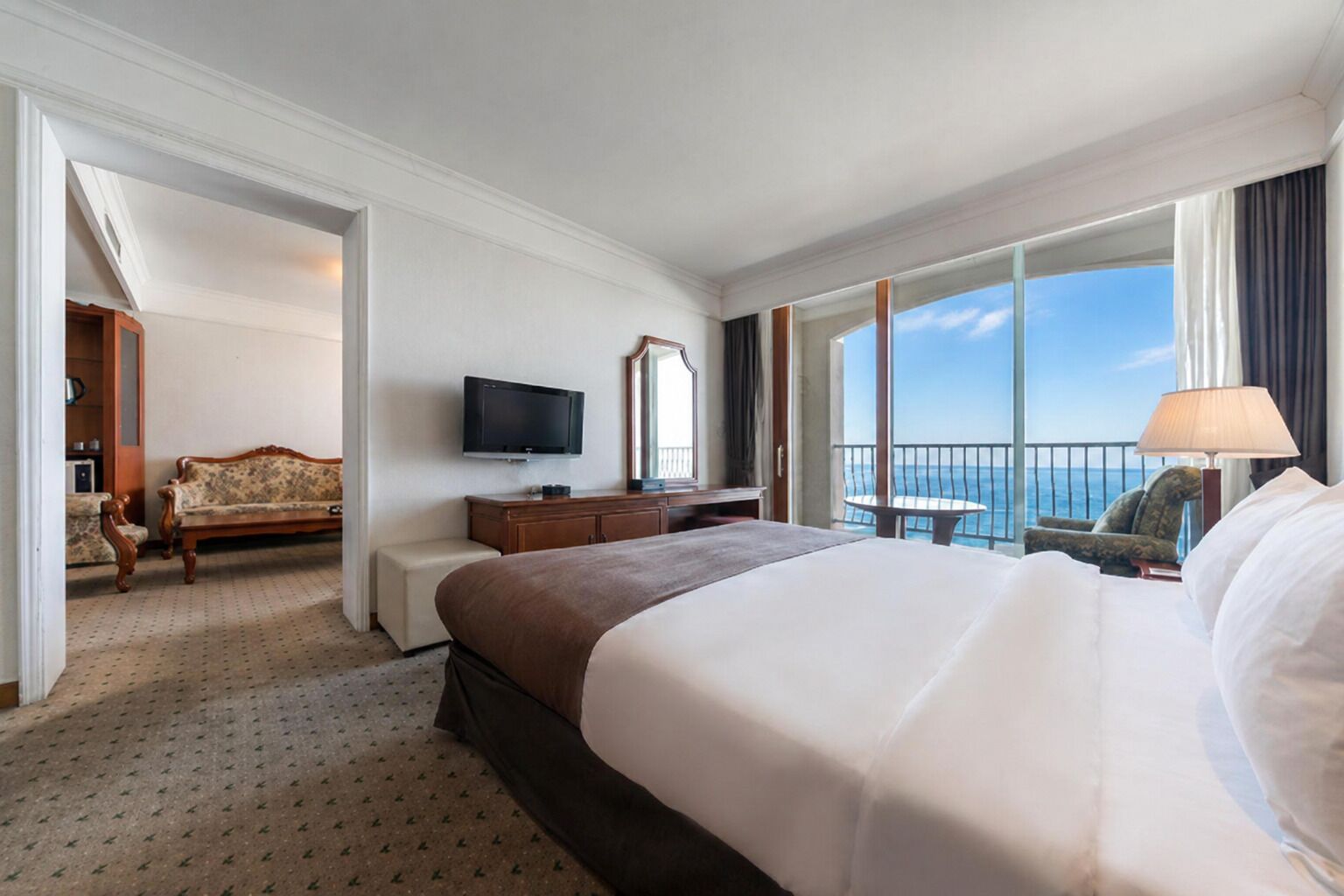 A hotel room with a large bed, a TV, a desk, a chair, and a balcony with a view of the sea.