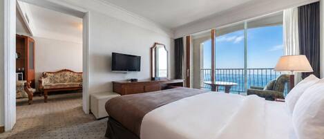 A hotel room with a large bed, a TV, a desk, a chair, and a balcony with a view of the sea.