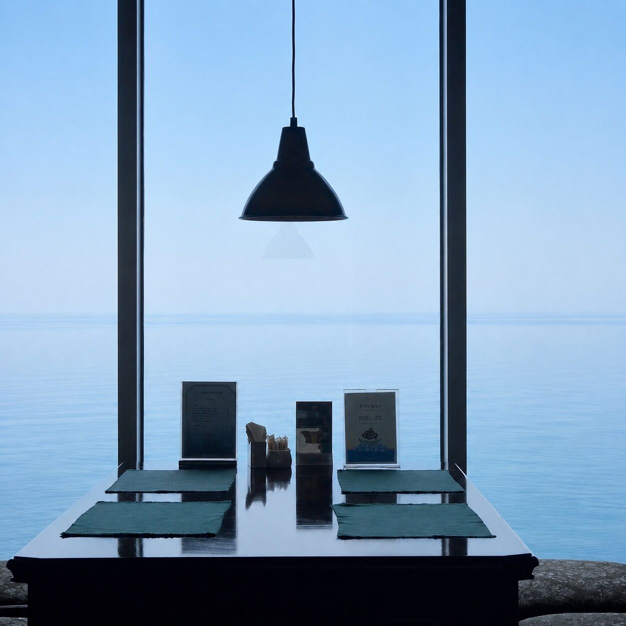 A table with a pendant lamp, two framed items, and a view of the sea through large windows.