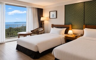 A hotel room with two beds, a chair, a table, a lamp, and a view of the sea.