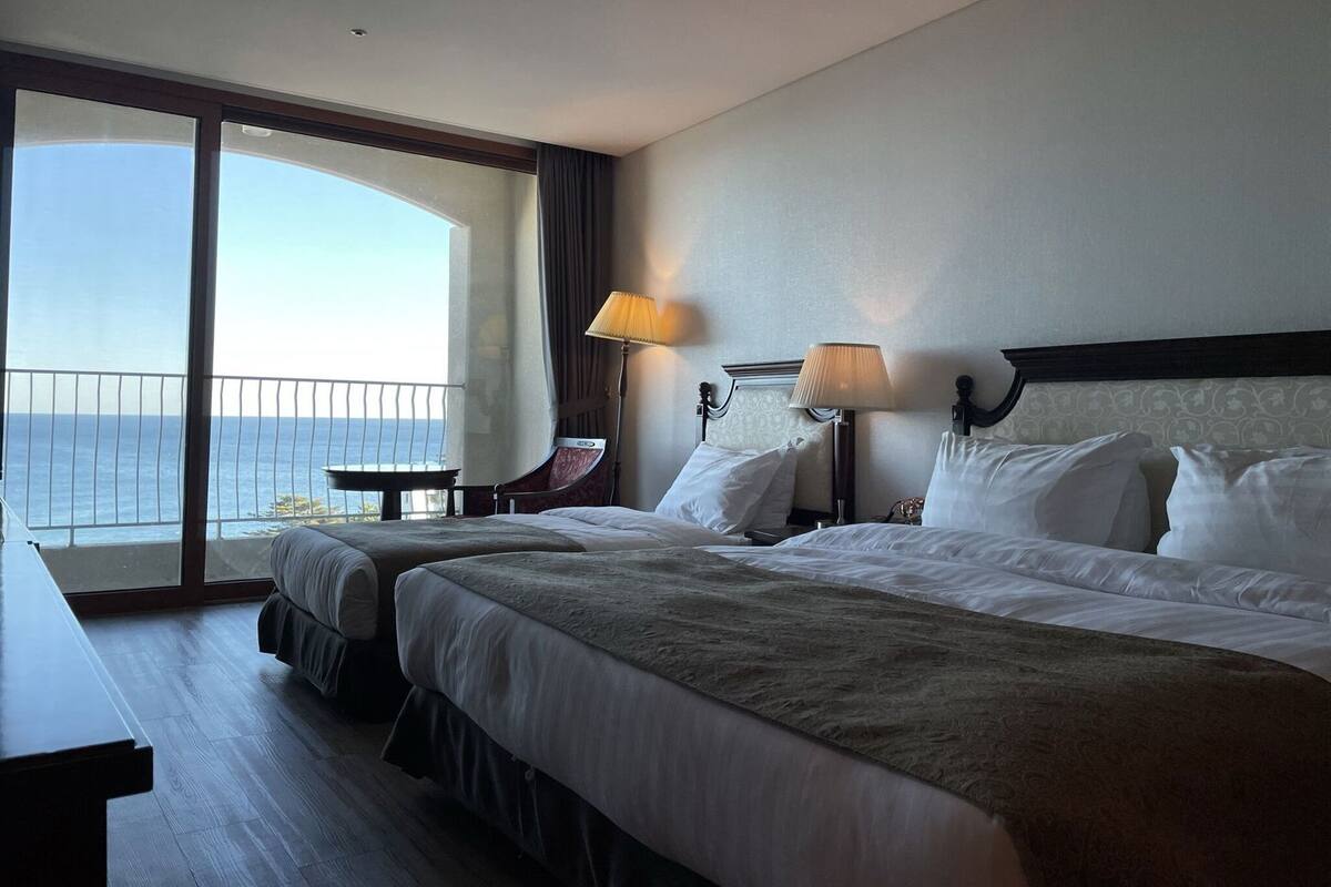 Ein Hotelzimmer mit einem großen Bett, einem kleineren Bett, einem Schreibtisch, einem Stuhl, einer Lampe und Blick auf den Ozean.