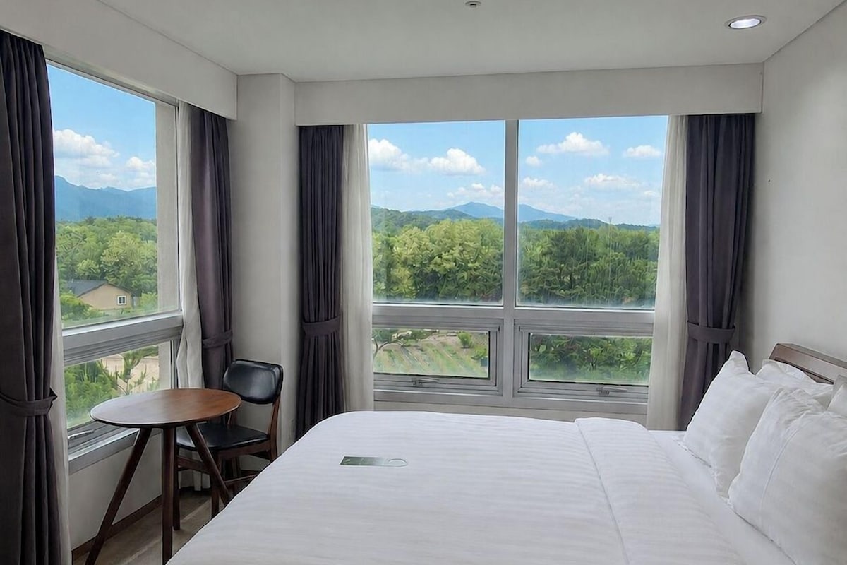 Une chambre d’hôtel avec un grand lit, une petite table et une chaise. Il y a une fenêtre qui donne sur un paysage verdoyant et montagneux.
