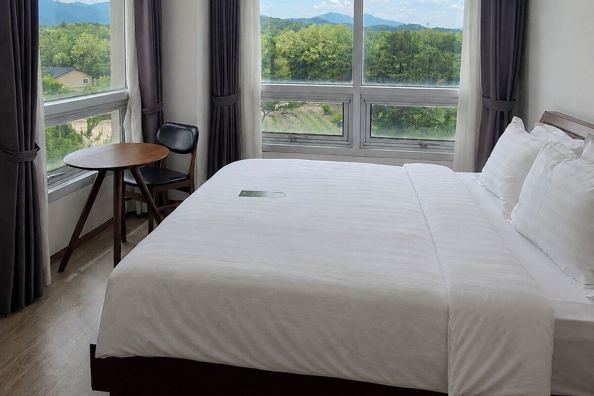 Une chambre d’hôtel avec un lit, une petite table, une chaise et une grande fenêtre donnant sur un paysage verdoyant et des montagnes.