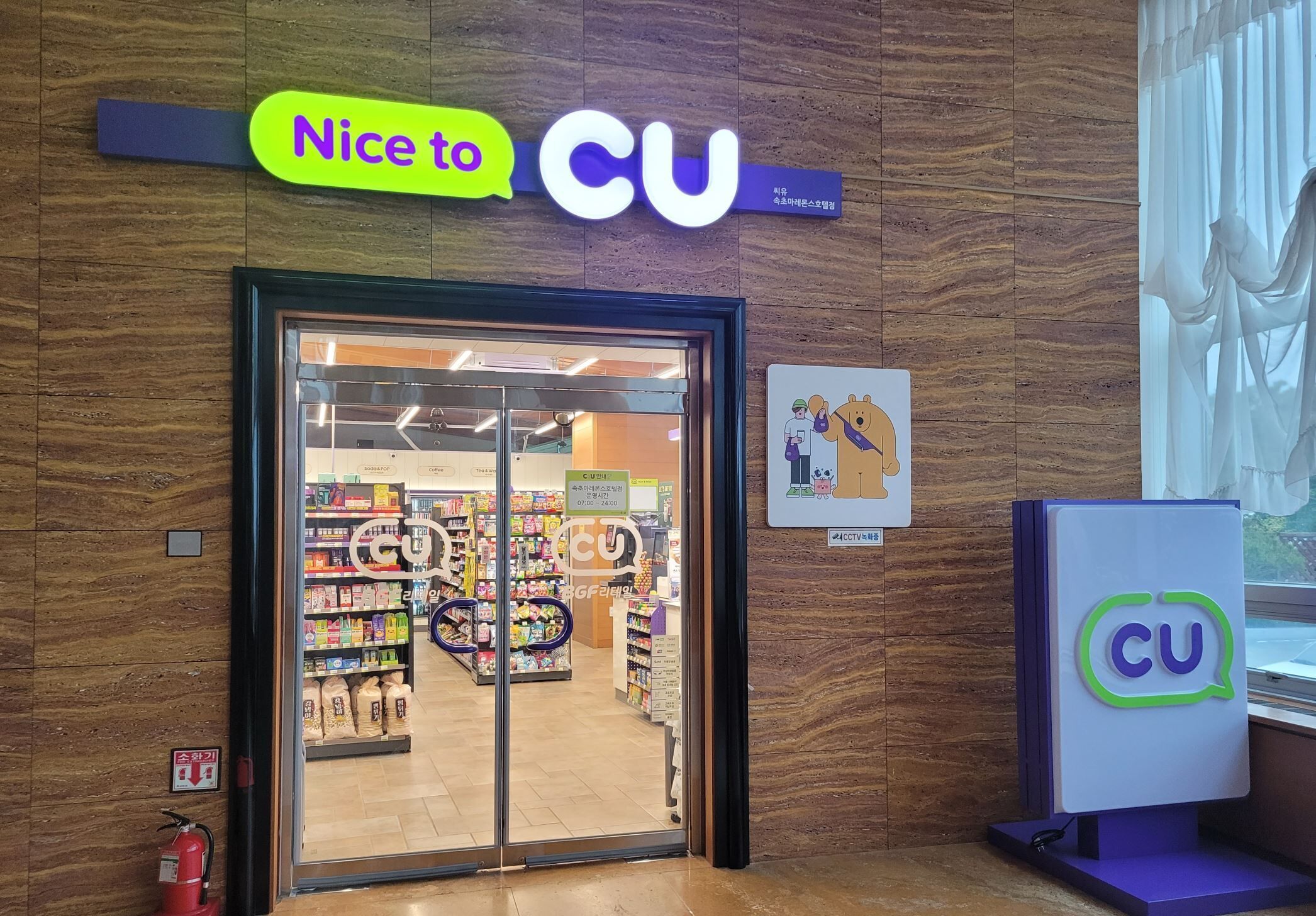 A CU store entrance with a sign above the door, a fire extinguisher on the wall, and a CU logo on a display stand.