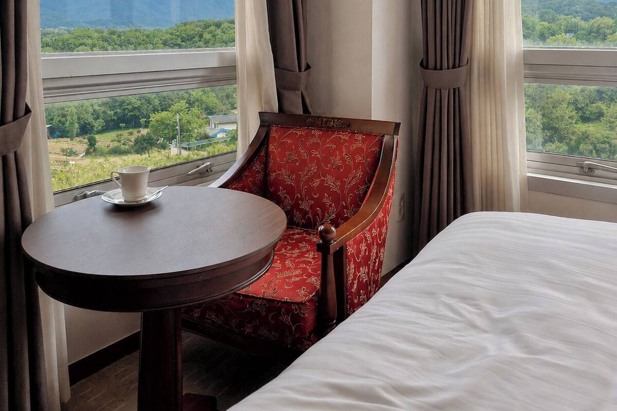 Ein Hotelzimmer mit einem Bett, einem runden Tisch mit Tasse, einem roten, gepolsterten Sessel und Blick auf Berge und Grün durch große Fenster.