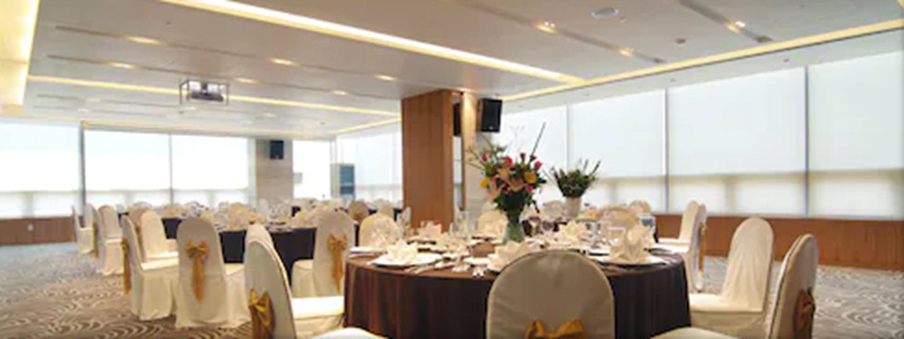 A banquet hall with round tables set for a formal event, featuring white chairs with gold sashes, a central floral arrangement, and a carpeted floor.