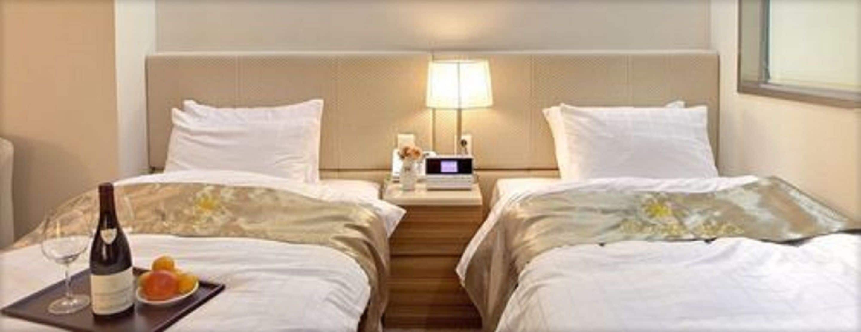 A hotel room with two beds, a bedside table with a lamp, a bottle of wine, and a plate of fruit.